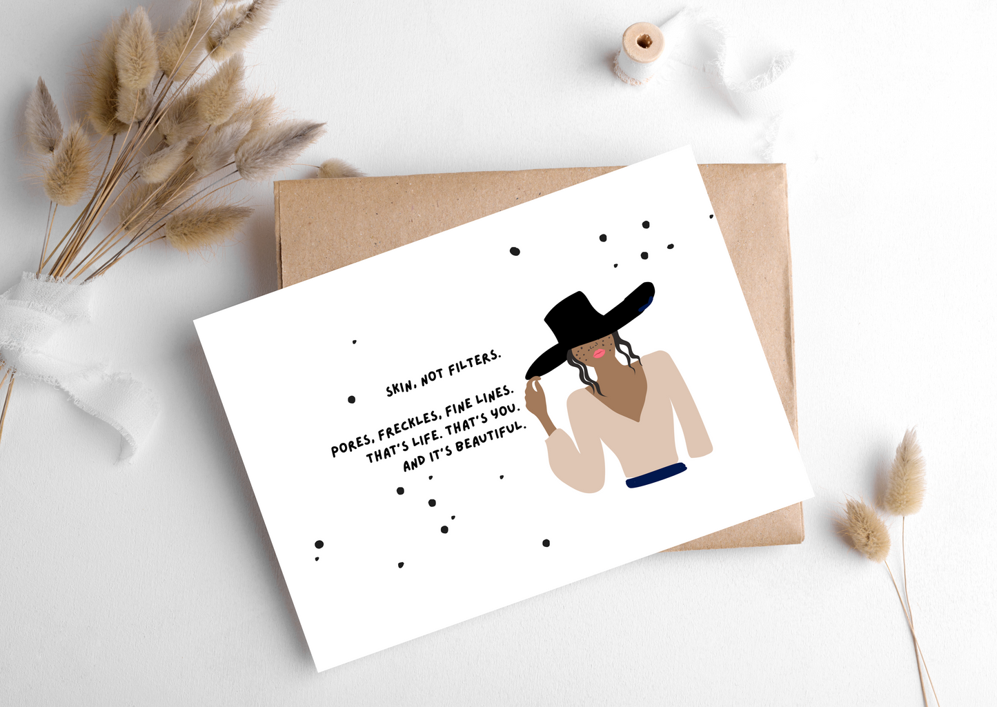 Card with illustration of a woman wearing a hat and text, placed on a white surface with dried plants.